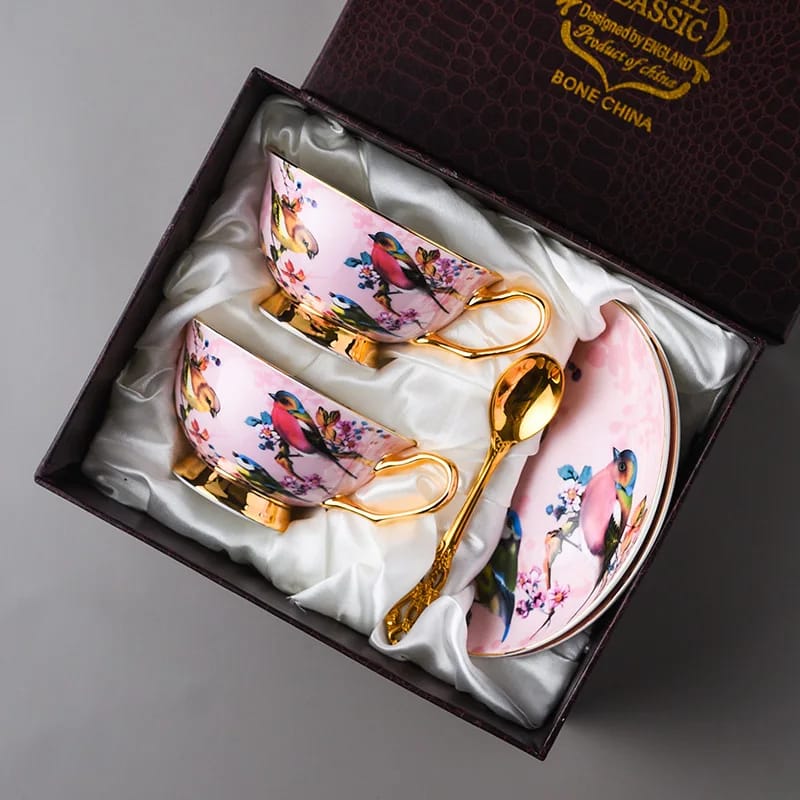Two pink cups decorated with birds and gold-tone handles, shown with a saucer and a gold-tone spoon